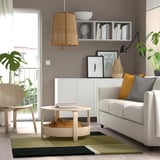 Modern living room with green/black rug, gray sofa, orange pillows, wooden coffee table, and wicker lamp.