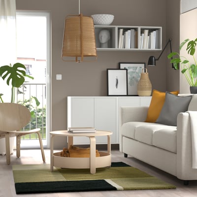 Modern living room with green/black rug, gray sofa, orange pillows, wooden coffee table, and wicker lamp.
