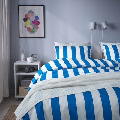 Modern bedroom with blue walls, art, nightstand with lamp, books, and blue-striped bedding.