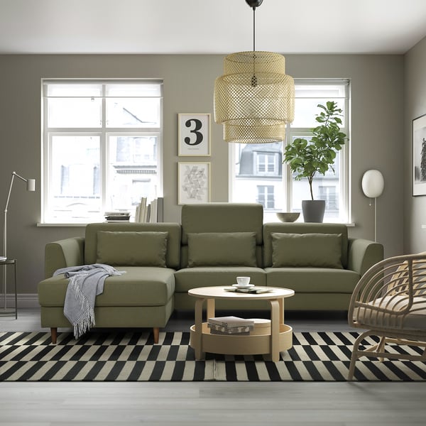 Modern living room with olive sofa, large windows, striped rug, coffee table, plant, wicker chair, and tiered light.