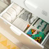Open white nursery drawers, displaying nappies, cloth wipes, and colorful toys neatly compartmentalized.