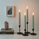 Four lit green-tinted candles on black holders, placed on a table with a picture frame and stacked books beside them.