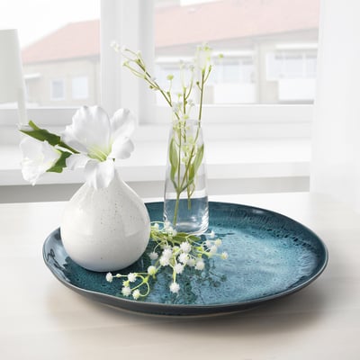 Two vases with white flowers on blue plate near window.