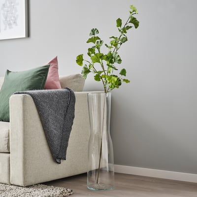 Vase named SMYCKA with green plant near beige sofa. Tall, clear glass, containing lifelike artificial greenery. Decorative, modern home decor piece.