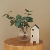 A small potted plant in a glass vase sits on a wooden stump, with an artificial stem, suitable for both indoor and outdoor use.