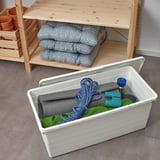Open plastic box with green cloth, rope, and water bottle. Near shelf with cushions and plants.