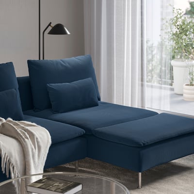 Blue sectional sofa with chaise, numerous pillows, modern lamp, and large window. Cosy living space.