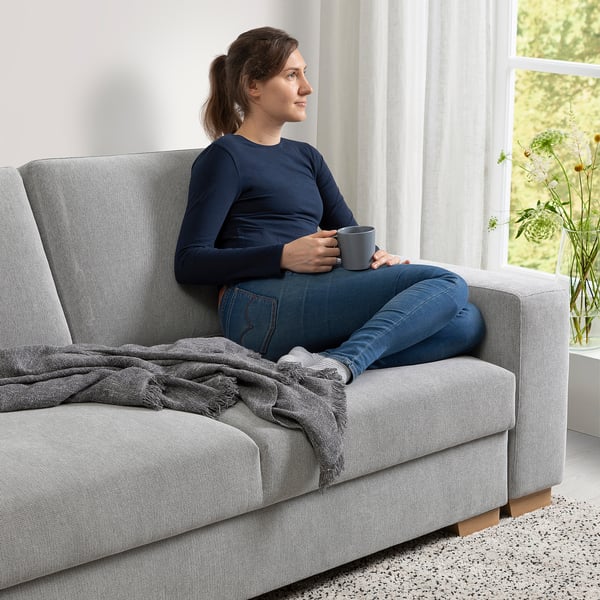 Woman sitting on a gray SÖRVALLEN sofa, holding a mug, with a blanket draped over the armrest. By a window with sheer curtains.