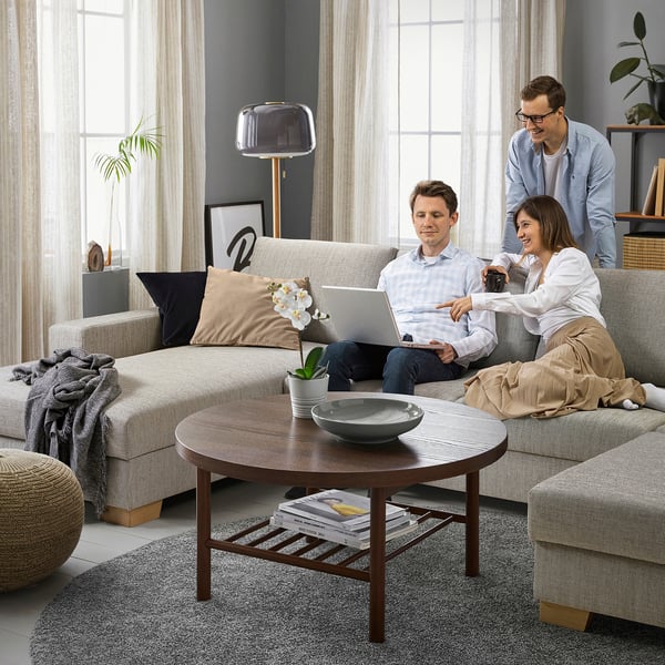 Three people in living room with sofa, table, plants; one works on laptop, others chat.