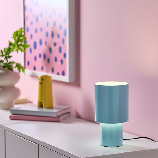 Modern turquoise SPETSBOJ lamp sits on a table, smaller than books, emits soft light.