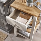 Wooden kitchen trolley with open drawer, holding kitchen towels, wooden cutting board, and tongs.