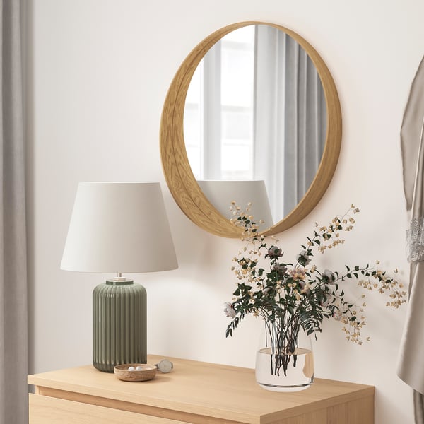 Minimalist room decor: round wooden mirror, lamp, and white vase with dried flowers on wooden table.