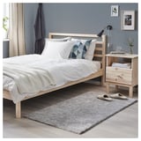 Modern bedroom with light wood bed, white bedding, and decorative rug. Grey and white decor, including wall art and plants.
