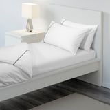 A neatly made bed with white sheets and pillows, elastic edges visible. A nightstand with a lamp beside it.