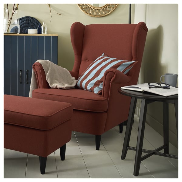 Red fabric wingback chair with a matching ottoman, striped pillow, and side table with a mug and book.