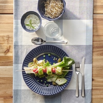 A wooden table with a striped placemat holds a blue plate of vegetable skewers, bowls of sauce and sprouts, a glass, and cutlery.