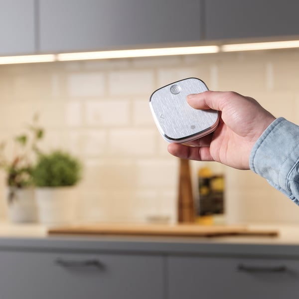 Hand holding STYRBAR remote in kitchen, controlling smart lights. Brushed steel finish, simple design for easy use.