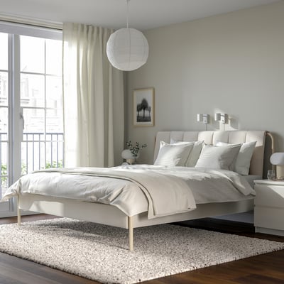 Modern bedroom with TÄLLÅSEN bed, light beige, grey wall, photo, curtains, rug, lamp, flowers.