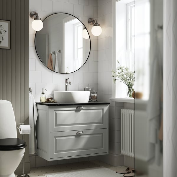Modern gray TÄNNFORSEN vanity with sink, mirror, lights, and drawers, essentials and vase; partial toilet.
