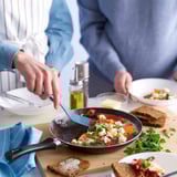 Two people serve a colorful, garnished dish from a non-stick frying pan on a wooden board with bread and condiments.