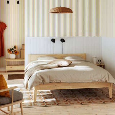 Modern bedroom with TAXÅSKLINT bed, pendant lights, striped wallpaper, neutral bedding, side table, rug, orange accent, and wooden bench with flowers.