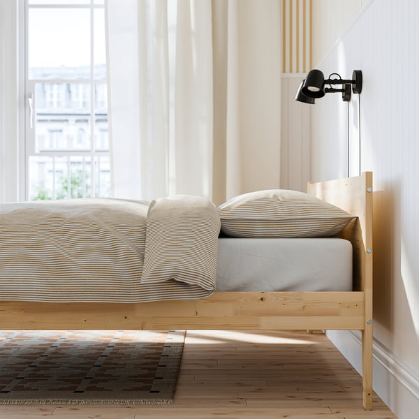 Bedroom with wooden bed frame, striped pillows, and wall lamp. Bed has clean, scandinavian design and wedge-shaped footboard.