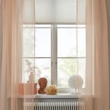 Pale orange curtains on tall window, sheer fabric diffuses light, decorative bust and vase on sill.