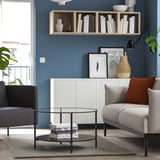 Round glass coffee table, black legs, central stack, grey rug, modern living room with grey sofa, orange pillow, blue wall, wooden floor.