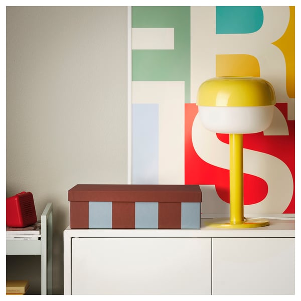Bright yellow lamp with white top on white cabinet, red speaker, next to brown box with blue stripes against colorful wall pattern.