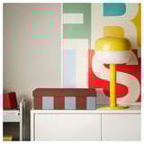 Bright yellow lamp with white top on white cabinet, red speaker, next to brown box with blue stripes against colorful wall pattern.