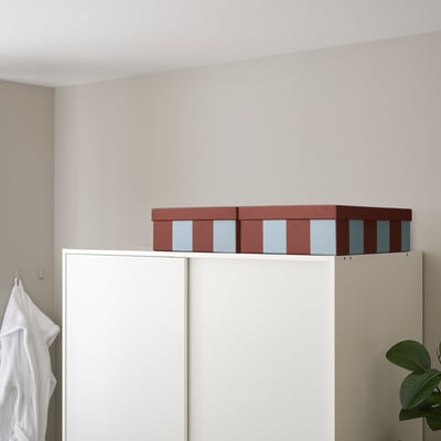 Two TJENA boxes, maroon with blue stripes, sit atop a white cabinet, larger than the white towel hanging nearby.