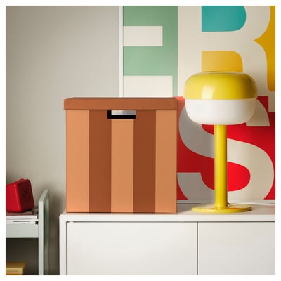 Bright yellow lamp with a spherical shade sits beside a brown TJENA storage box on a white cabinet, against a colorful geometric wallpaper.