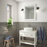 Modern bathroom with TOFTBYN white mirror above white vanity. Gray tiled walls, hanging blue towels, and potted plant.