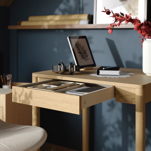 Desk with open drawer, holding stationery, tablet, and headphones. Shelf above with books, vase, and blossoms.