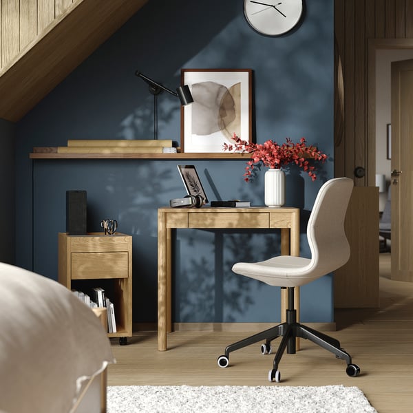 Modern home office with light wood desk, white chair, laptop, lamp, books, and red flowers.