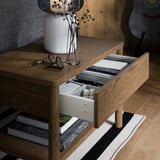 Wooden side table with open shelf and soft-close drawer, holding books, dice, and a vase with dried flowers.