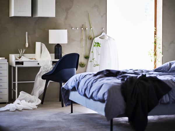 Modern bedroom with blue comforter on bed, blue chair, desk with lamp, and hanging plants.