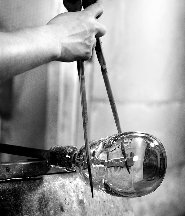 Black and white hands shaping molten glass with tools.