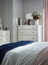 Cozy bedroom with white dressers. One has a glass vase with flowers. Blue blanket on bed.