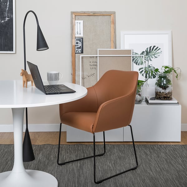 Brown leather TOSSBERG chair at white desk with laptop, mug, and horse. White shelf behind with art, plant, and books.