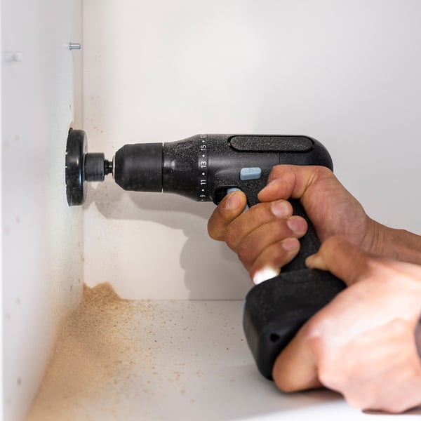 A hand uses a TRIXIG drill to make a hole in a white surface, creating dust.