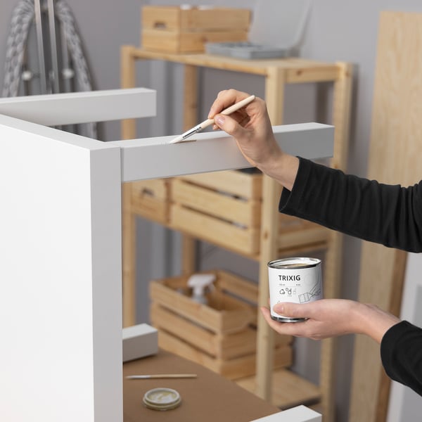 Person painting white furniture with a brush using TRIXIG paint tin. Wooden shelves and tools in background.
