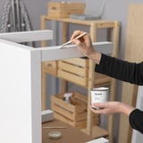 Person painting white furniture with a brush using TRIXIG paint tin. Wooden shelves and tools in background.