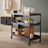Modern metal three-shelf trolley with wheels, displaying mugs, apples, and bags.