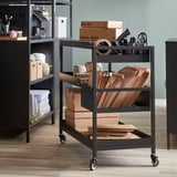 Black metal trolley with wheels, holding mail boxes, tape, and files, next to a shelving unit.