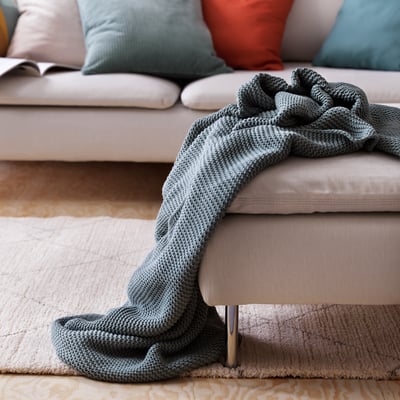 A cozy living room with a light grey sofa, adorned with a soft, gray knitted blanket casually draped over one arm.