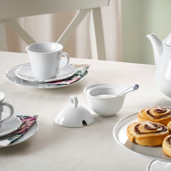 Table set with porcelain cups, plates, teapot, sugar bowl, pastries, and floral napkins on a beige cloth.
