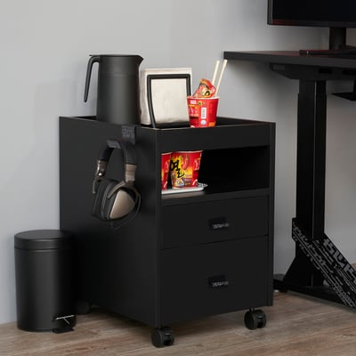 Black UPPSPEL mobile storage unit with two drawers, holding headphones, drinks, snacks, and a laptop, next to a standing desk.