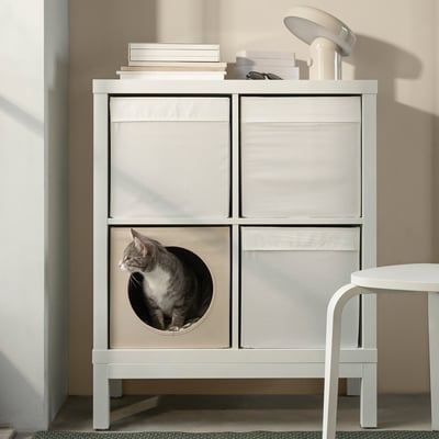 White UTSÅDD cat house in kallax shelf, cat peeking through circular cutout. Books and lamp on top, stool to right.