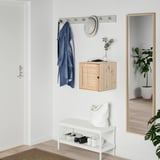 Small entryway setup with VÄLJARE cabinet, featuring light wood and woven texture, hanging blue coat, white hat, and small plants.